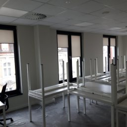 Rollcom Łukasz Feliks - Wnętrze biura z trzema oknami z częściowo zasłoniętymi oknami, liczne białe biurka z metalowymi nogami, krzesło biurowe z lewej strony, sufit kasetonowy z okrągłymi nawiewami, szara wykładzina...