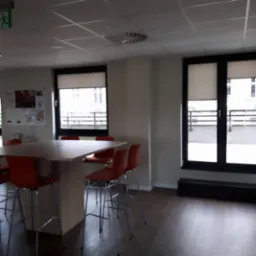 Wnętrze biurowe z dwoma oknami z roletami zwijanymi, widoczne krzesła barowe i duży stół konferencyjny.