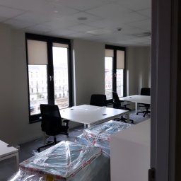 Rollcom Łukasz Feliks - Wnętrze biura z jasnymi ścianami i podwieszanym sufitem, dwa okna z częściowo opuszczonymi, jasnymi osłonami, biurka z czarnymi krzesłami, w tle widok na budynek i tory kolejowe, w pierwszym planie...