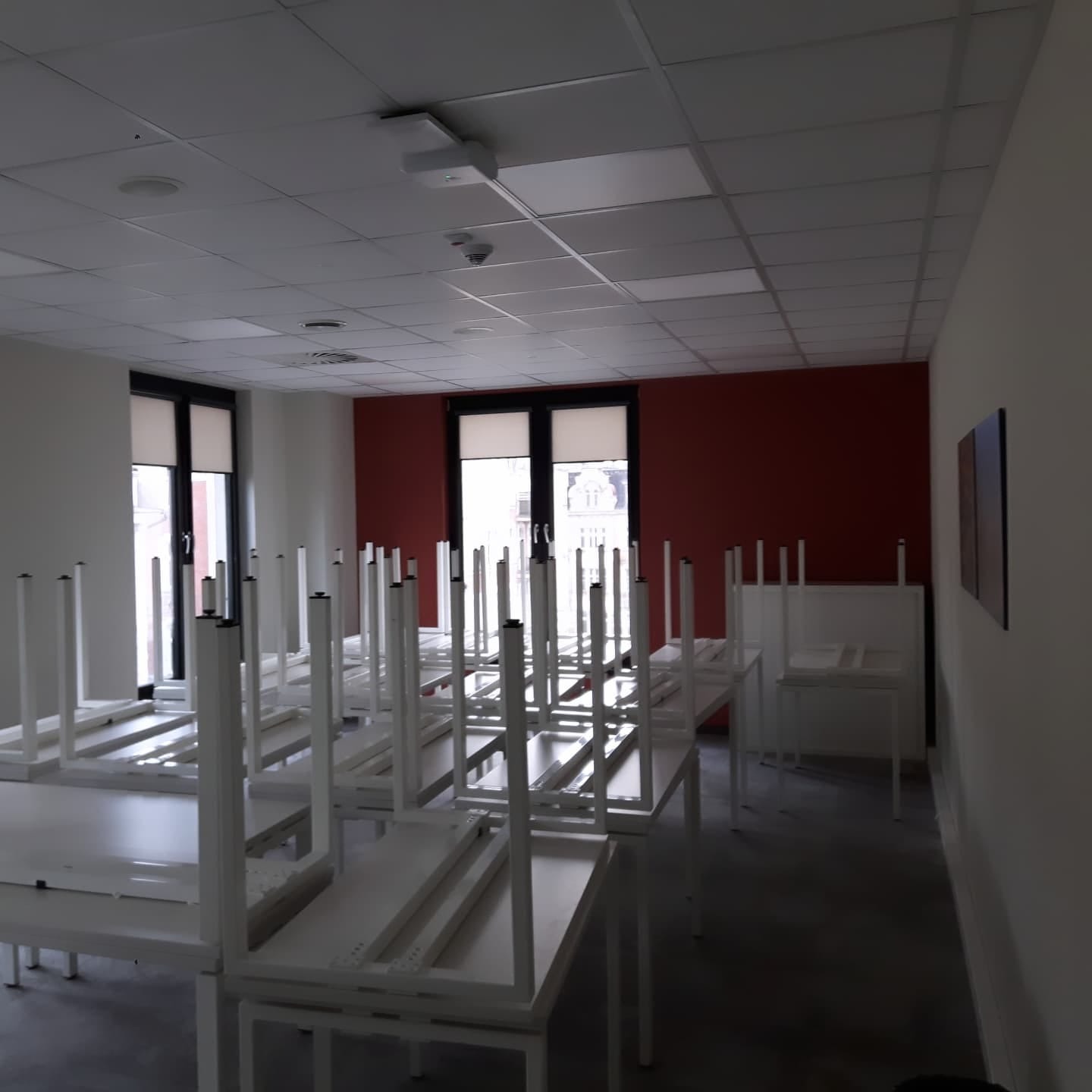 Sala konferencyjna z białymi, częściowo rozłożonymi stołami, widoczne okna z zasłoniętymi oknami i czerwona ściana w tle