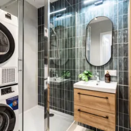 Nowoczesna łazienka z pralką i suszarką w kolumnie, kabiną prysznicową walk-in z ciemnymi, błyszczącymi płytkami oraz drewnianą szafką z umywalką i okrągłym lustrem.
