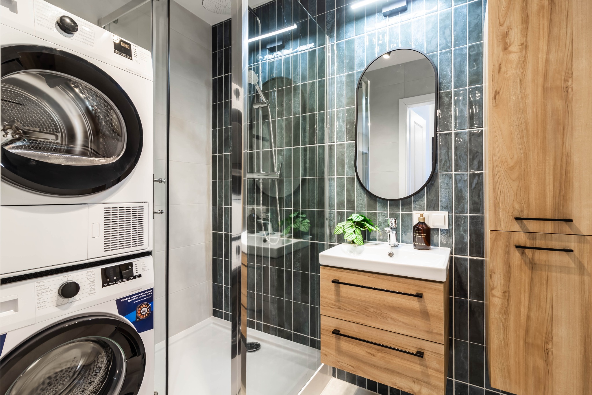 Nowoczesna łazienka z pralką i suszarką w kolumnie, kabiną prysznicową walk-in z ciemnymi, błyszczącymi płytkami oraz drewnianą szafką z umywalką i okrągłym lustrem.