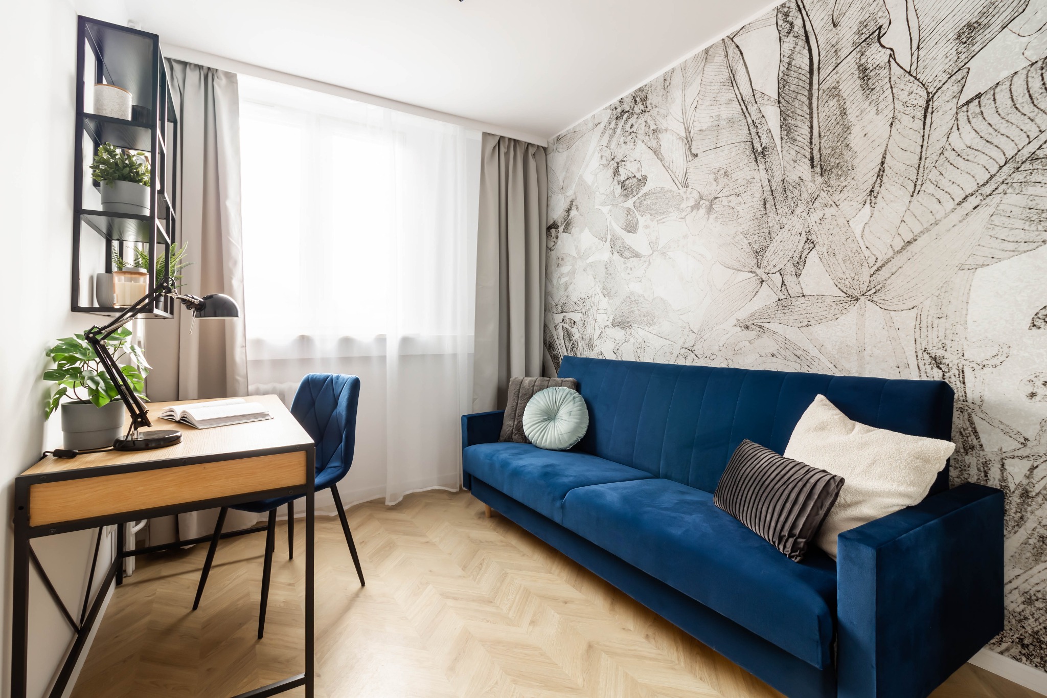 Pokój z niebieską sofą, biurkiem z lampką i ścianą z motywem roślinnym, jasne wnętrze z parkietem w jodełkę.