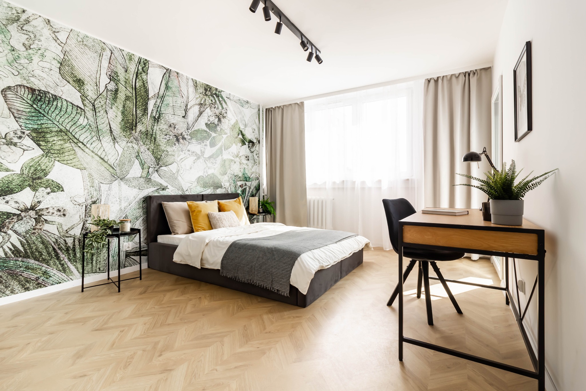 Jasna sypialnia z łóżkiem, biurkiem i ścianą z motywem roślinnym, parkiet w jodełkę na podłodze, oświetlenie punktowe na suficie.