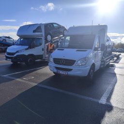 RBTrans Rafał Bobiński - Dwa białe samochody dostawcze typu laweta zaparkowane na parkingu. Jeden z nich, marki Mercedes, ma załadowany na platformie szary samochód osobowy. W tle widoczny pracownik w pomarańczowej...