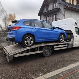 RBTrans Rafał Bobiński - Niebieskie BMW kombi z uszkodzonym przodem, zabezpieczone pasami na platformie lawety, na tle szarego domu z balkonami.