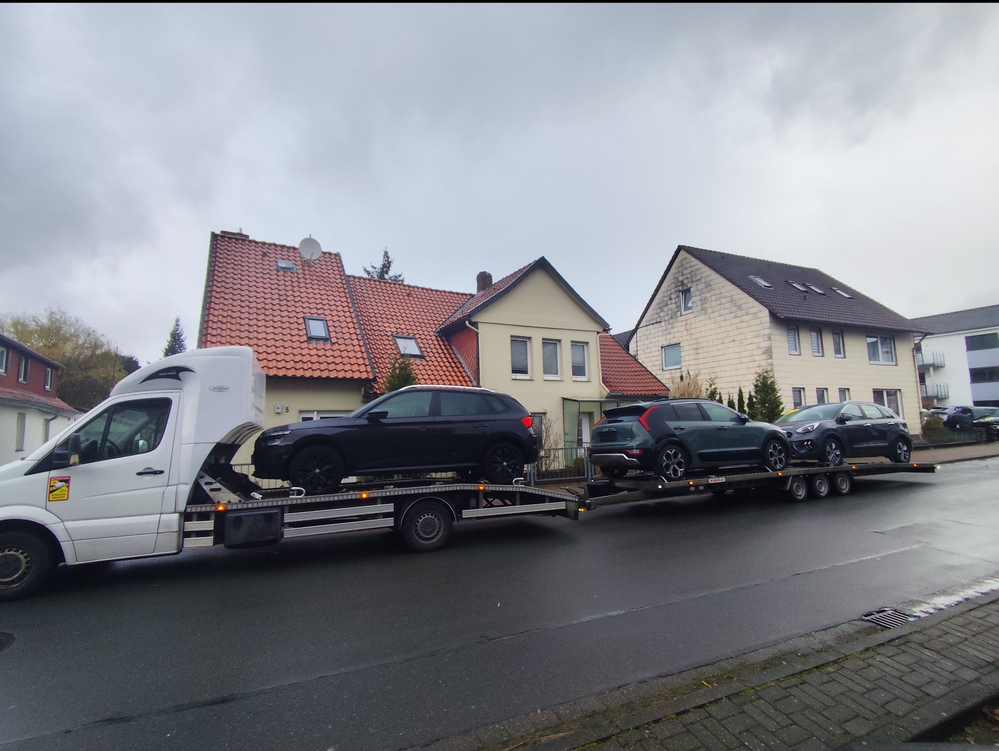 Biała laweta z trzema samochodami typu SUV na platformie transportowej, zaparkowana na mokrej ulicy przed domami w niemieckim miasteczku, pochmurny dzień.
