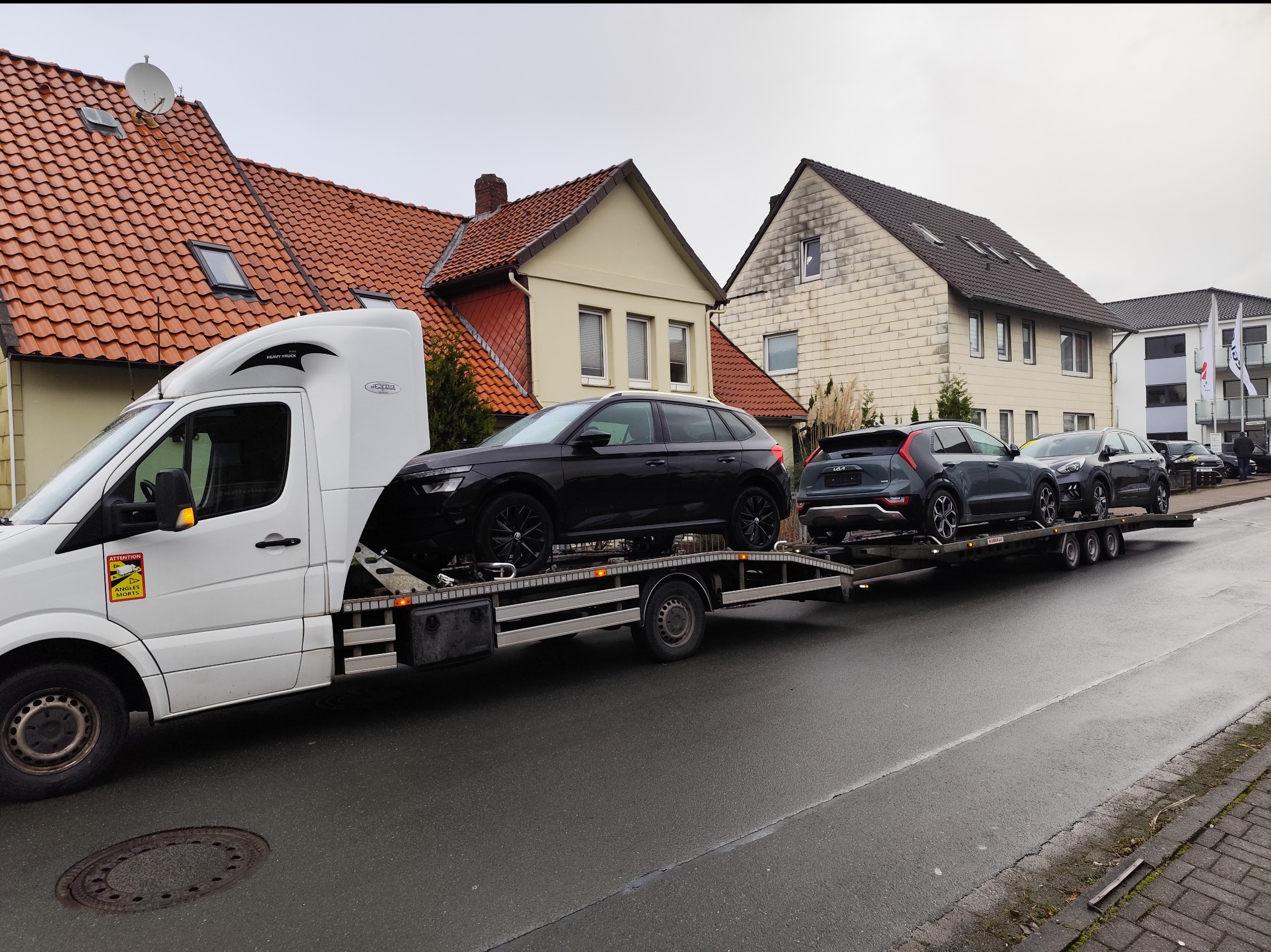 Biała laweta z trzema szarymi samochodami osobowymi marki Kia i Skoda na platformie, zaparkowana na ulicy przed budynkami mieszkalnymi w pochmurny dzień.