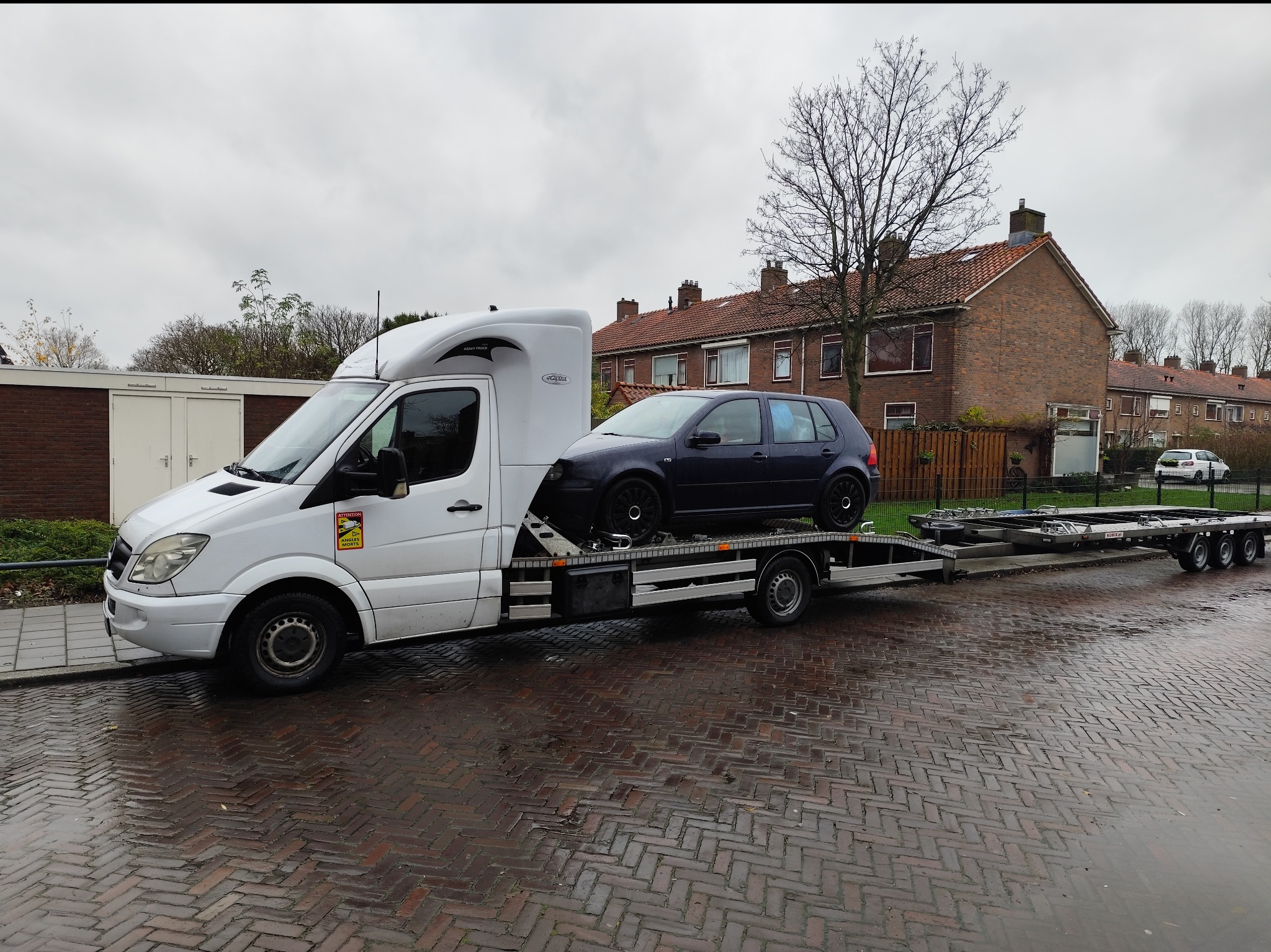 Biała laweta z ciemnym hatchbackiem na platformie, zaparkowana na mokrym brukowanym chodniku w europejskim mieście, widoczny znak 'Attention Angles Morts' na kabinie.