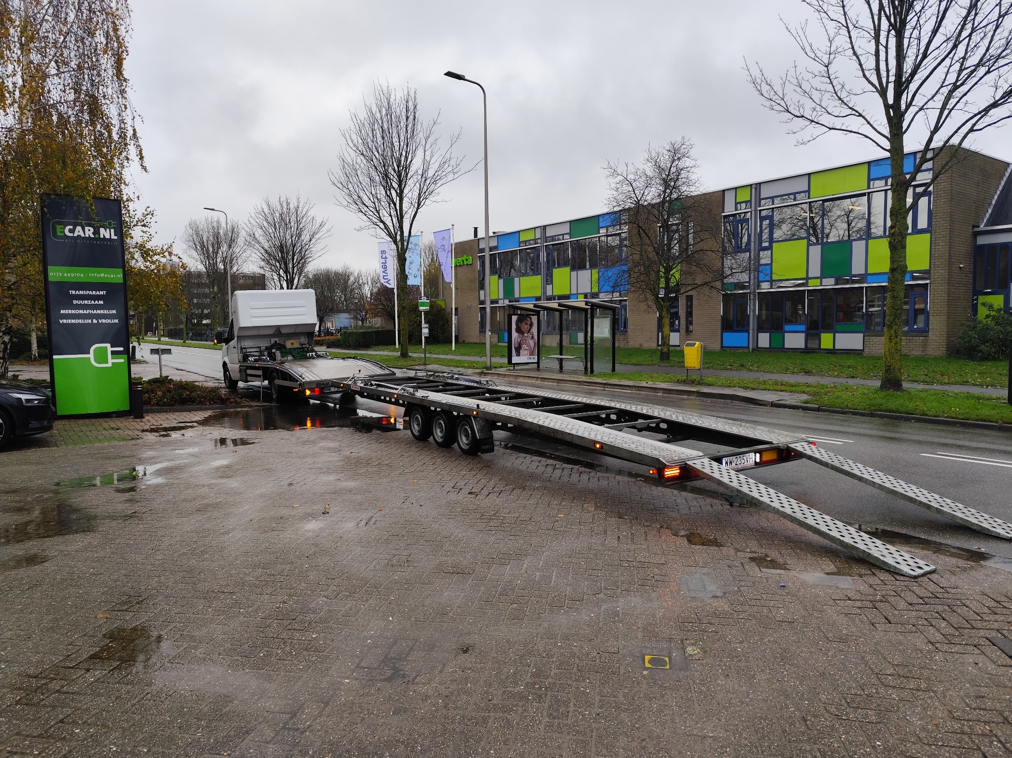 Laweta z rozłożonymi najazdami zaparkowana na mokrym bruku przed budynkiem z kolorowymi oknami i zielonym szyldem firmy Ecar.nl.