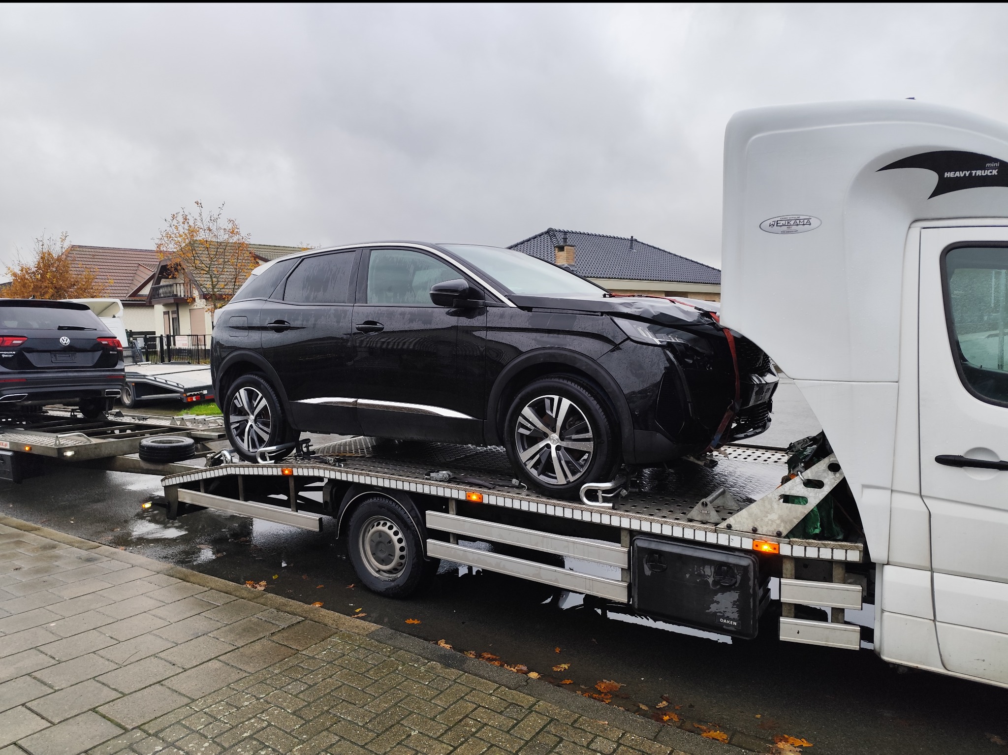 Czarny Peugeot 3008 z uszkodzonym przodem, zabezpieczony łańcuchami na platformie lawety, obok drugi samochód na lawecie, widok z boku na tle domów jednorodzinnych w pochmurny dzień.