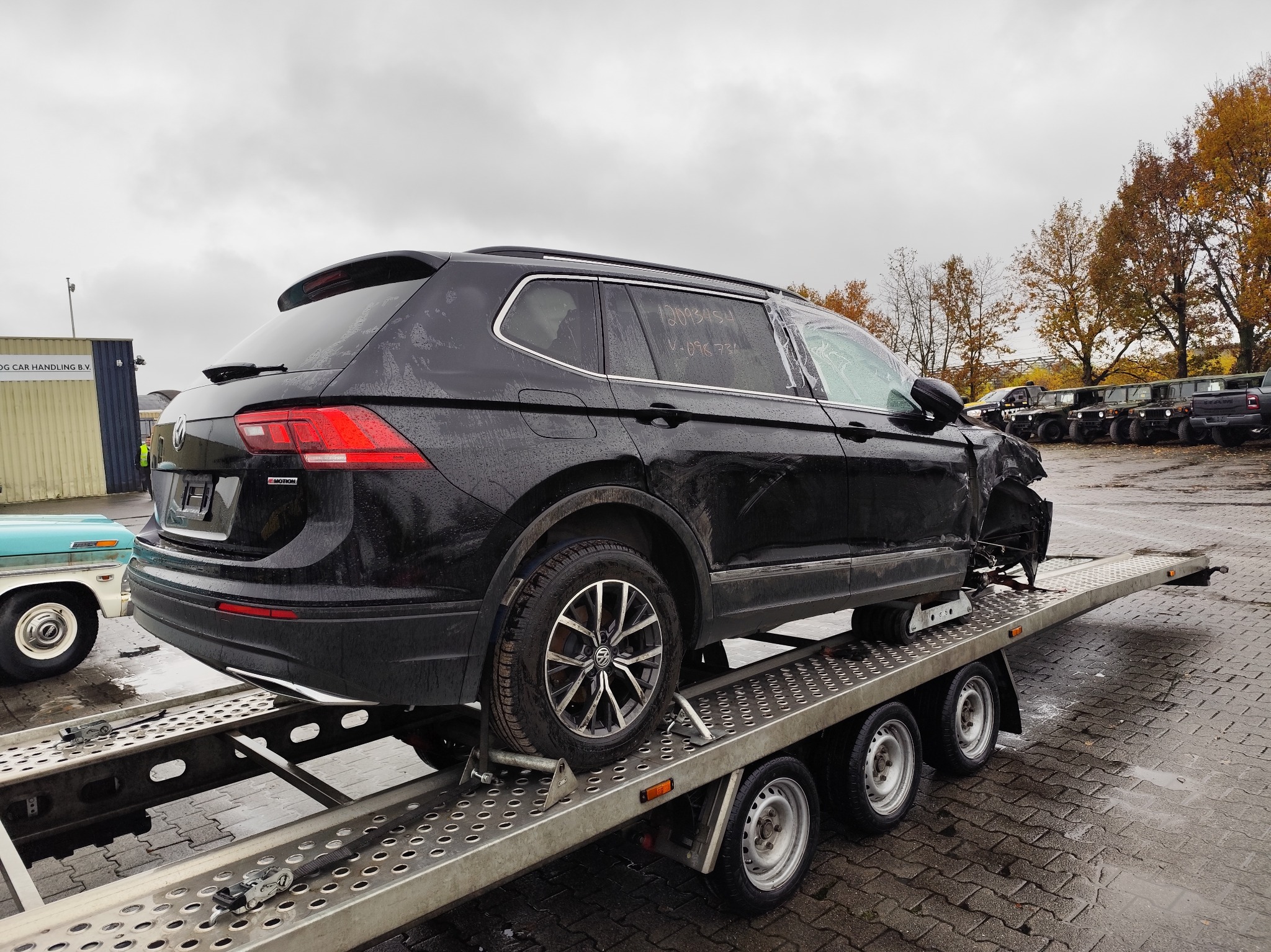 Uszkodzony, czarny Volkswagen Tiguan z napisem na szybie, transportowany na metalowej lawecie z trzema osiami, na mokrym placu postojowym z drzewami w tle.