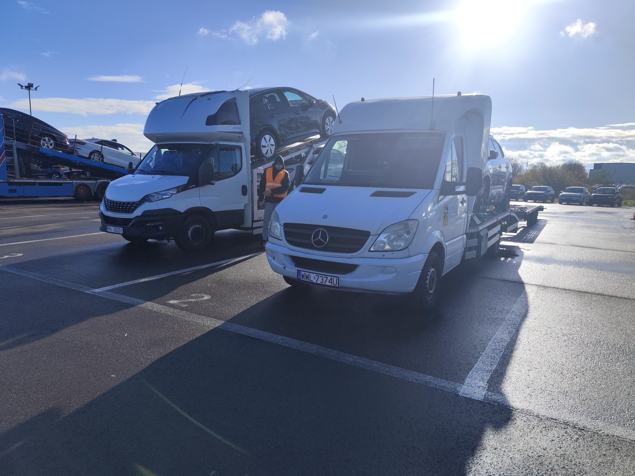 Dwa białe samochody dostawcze typu laweta zaparkowane na parkingu. Jeden z nich, marki Mercedes, ma załadowany na platformie szary samochód osobowy. W tle widoczny pracownik w pomarańczowej...