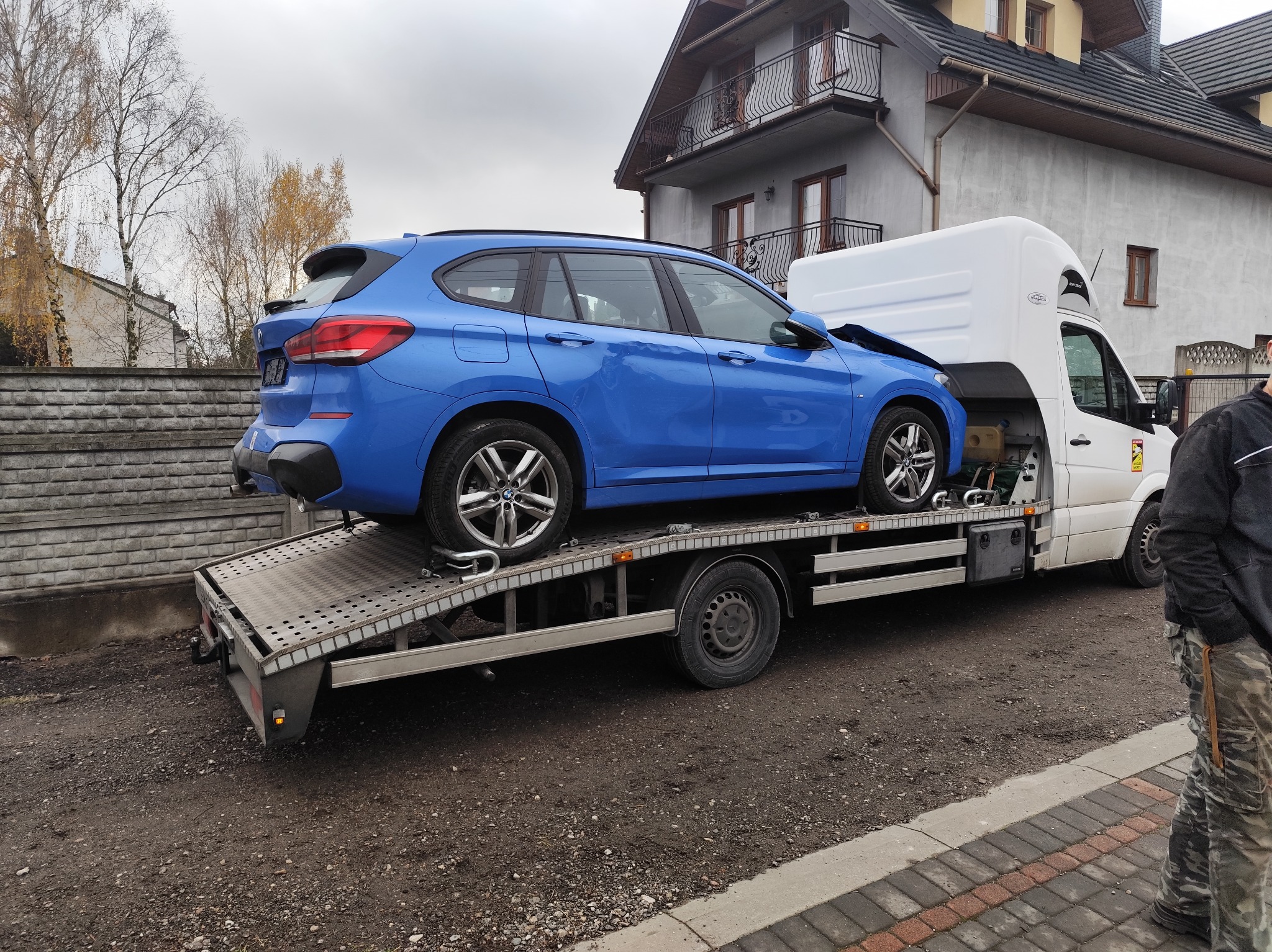 Niebieskie BMW kombi z uszkodzonym przodem, zabezpieczone pasami na platformie lawety, na tle szarego domu z balkonami.