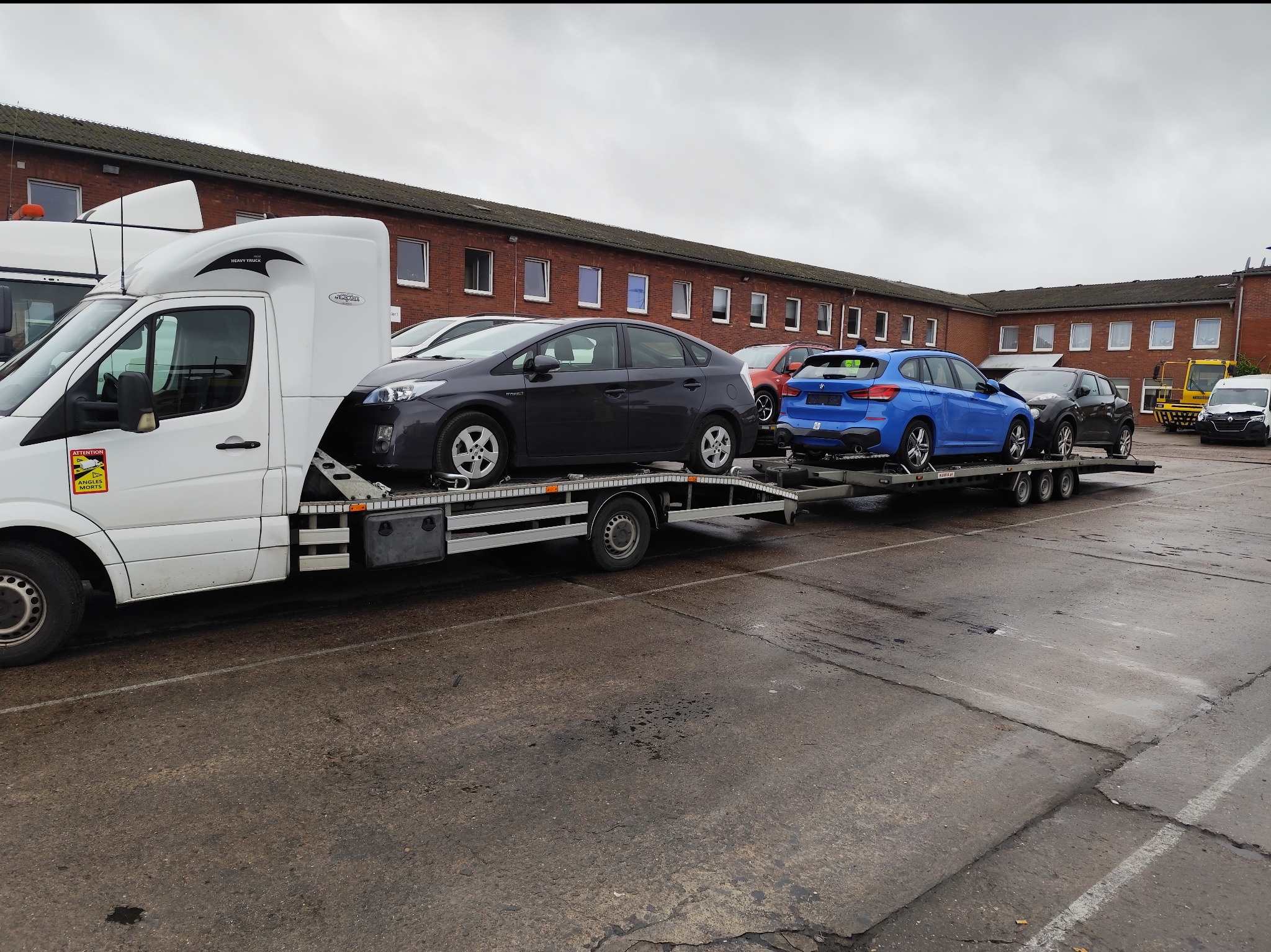 Laweta z trzema samochodami osobowymi różnych marek (Toyota Prius, BMW X1, Nissan Juke) na parkingu przed budynkiem z czerwonej cegły w pochmurny dzień.