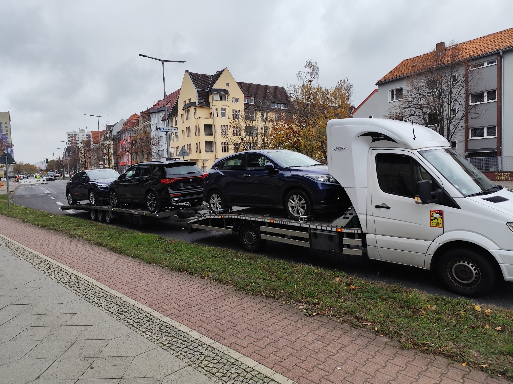Biała laweta z trzema samochodami osobowymi na platformie, zaparkowana na poboczu ulicy w zabudowie miejskiej, widoczna naklejka ostrzegawcza 'Attention Angles Morts'.