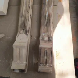 Dwie drewniane kolumny z dekoracyjnymi detalami, jedna oczyszczona, druga z widocznymi śladami zabrudzeń i starej farby, ukazujące postęp prac renowacyjnych.