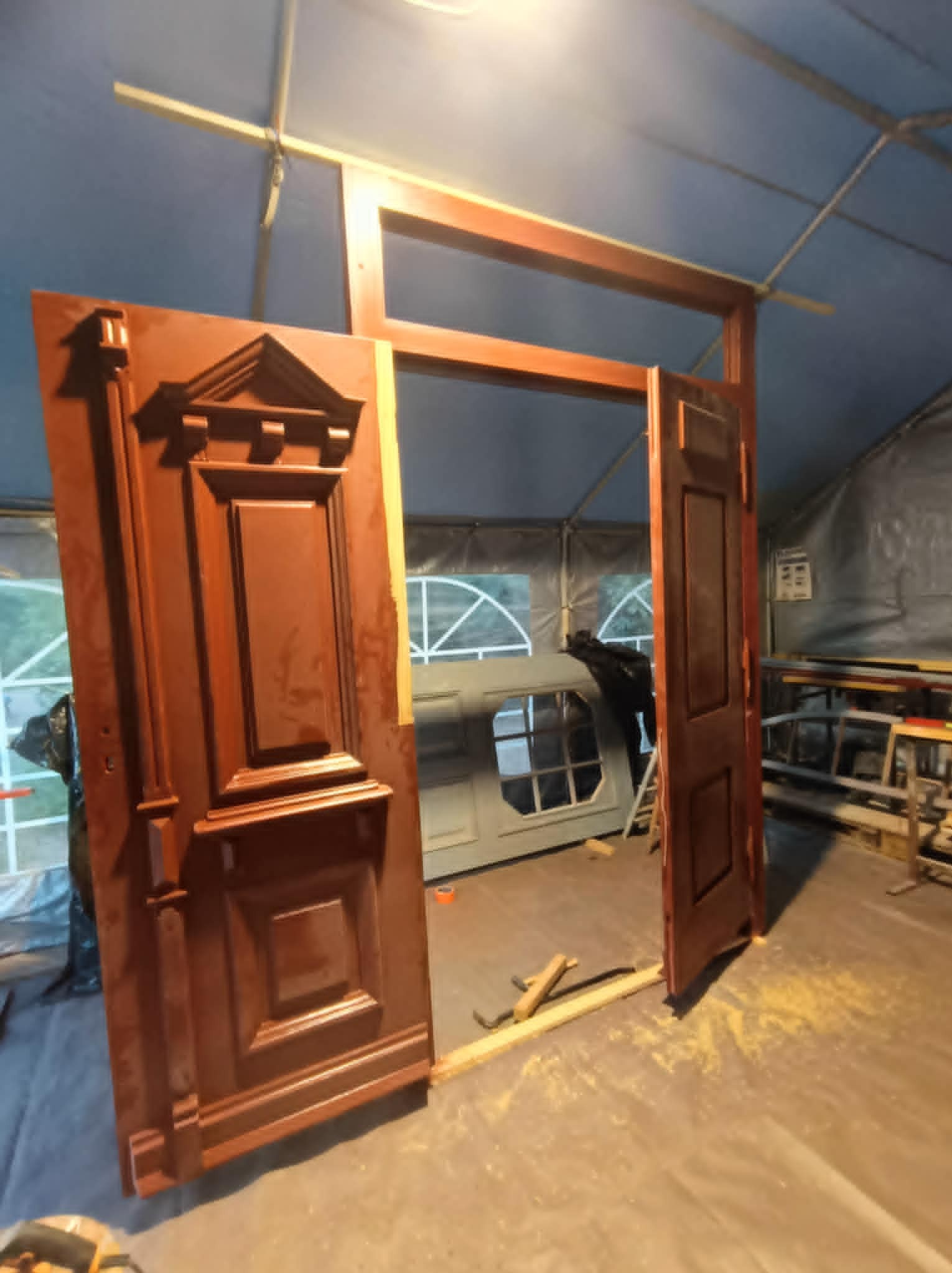 Odrestaurowane, brązowe, drewniane drzwi z dekoracyjnymi elementami, częściowo otwarte, w trakcie prac renowacyjnych wewnątrz namiotu ochronnego.