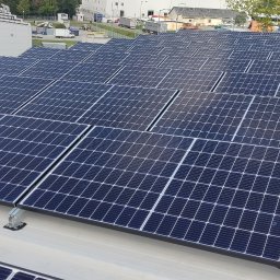 VIA - Rozległa instalacja paneli słonecznych na płaskim dachu budynku przemysłowego, widoczna drabina dostępu, w tle zaparkowane ciężarówki i budynki.