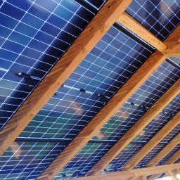 VIA - Widok od dołu na panele słoneczne, zainstalowane między drewnianymi belkami konstrukcji dachu, z widocznymi złączami kablowymi.