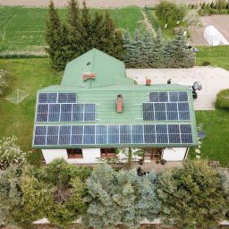 VIA - Widok z lotu ptaka na dom z zielonym dachem pokrytym panelami słonecznymi, otoczony zielenią i drzewami, z widocznym ogrodem i fragmentem drogi dojazdowej.