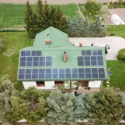 Widok z lotu ptaka na dom z zielonym dachem pokrytym panelami słonecznymi, otoczony zielenią i drzewami, z widocznym ogrodem i fragmentem drogi dojazdowej.