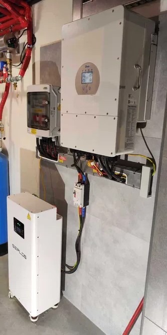 Kompletny system zasilania awaryjnego: biała bateria magazynu energii SEPLOS na kółkach, inwerter z wyświetlaczem i rozdzielnia elektryczna zamontowane na ścianie z szarych płytek, widoczne...