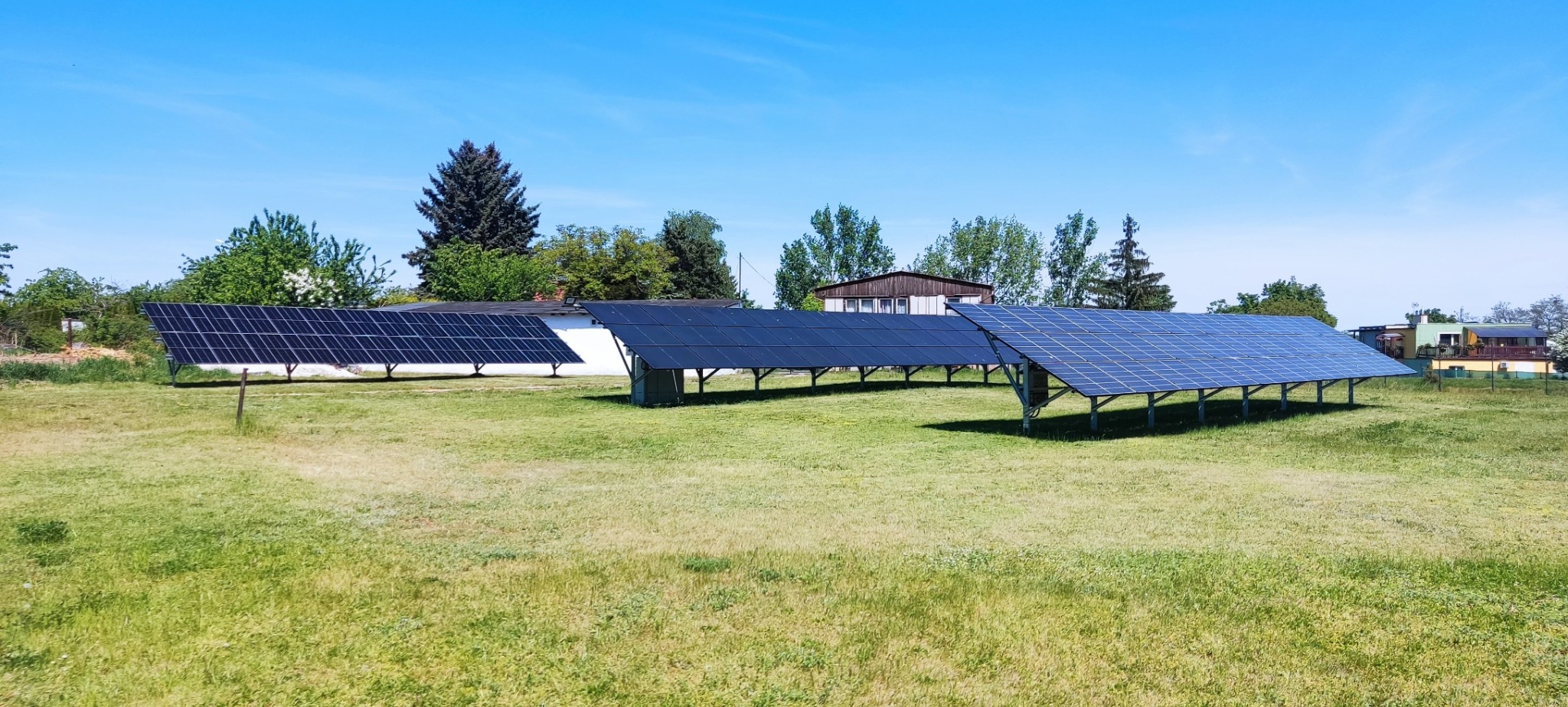 Rozległa instalacja fotowoltaiczna na gruncie, z panelami ustawionymi rzędami na metalowych konstrukcjach, na tle zielonego trawnika i budynków gospodarczych pod błękitnym niebem.