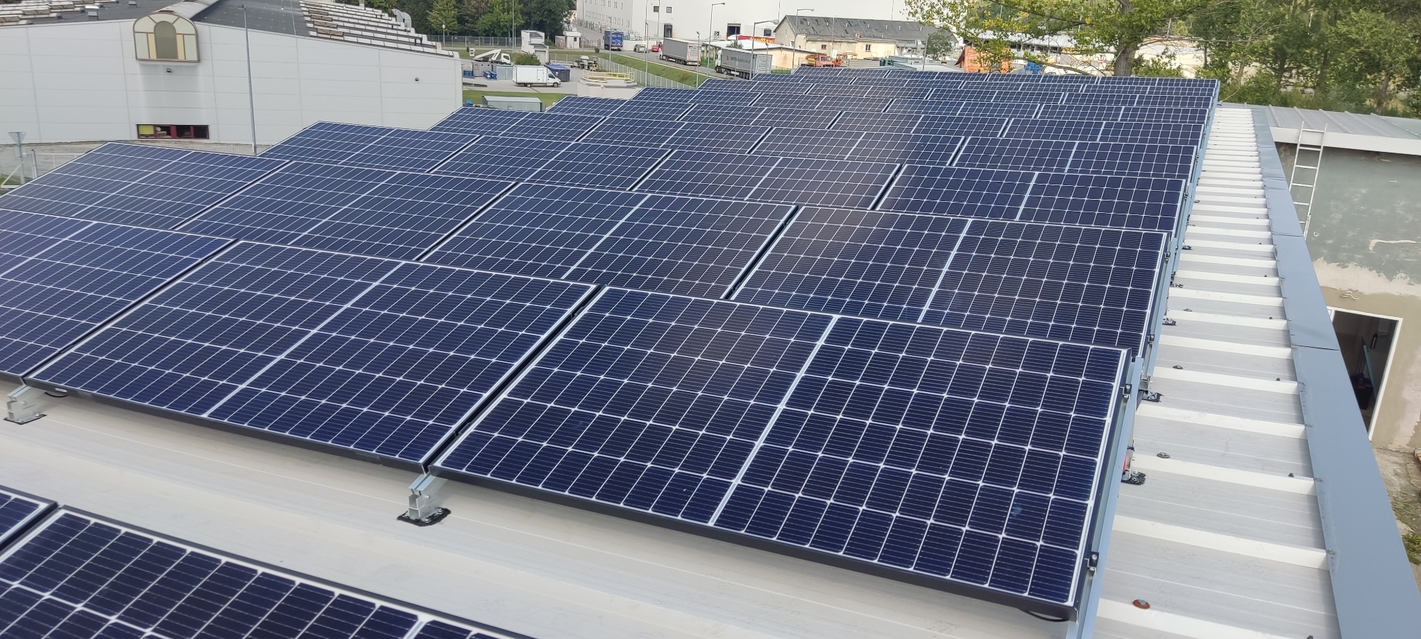 Rozległa instalacja paneli słonecznych na płaskim dachu budynku przemysłowego, widoczna drabina dostępu, w tle zaparkowane ciężarówki i budynki.