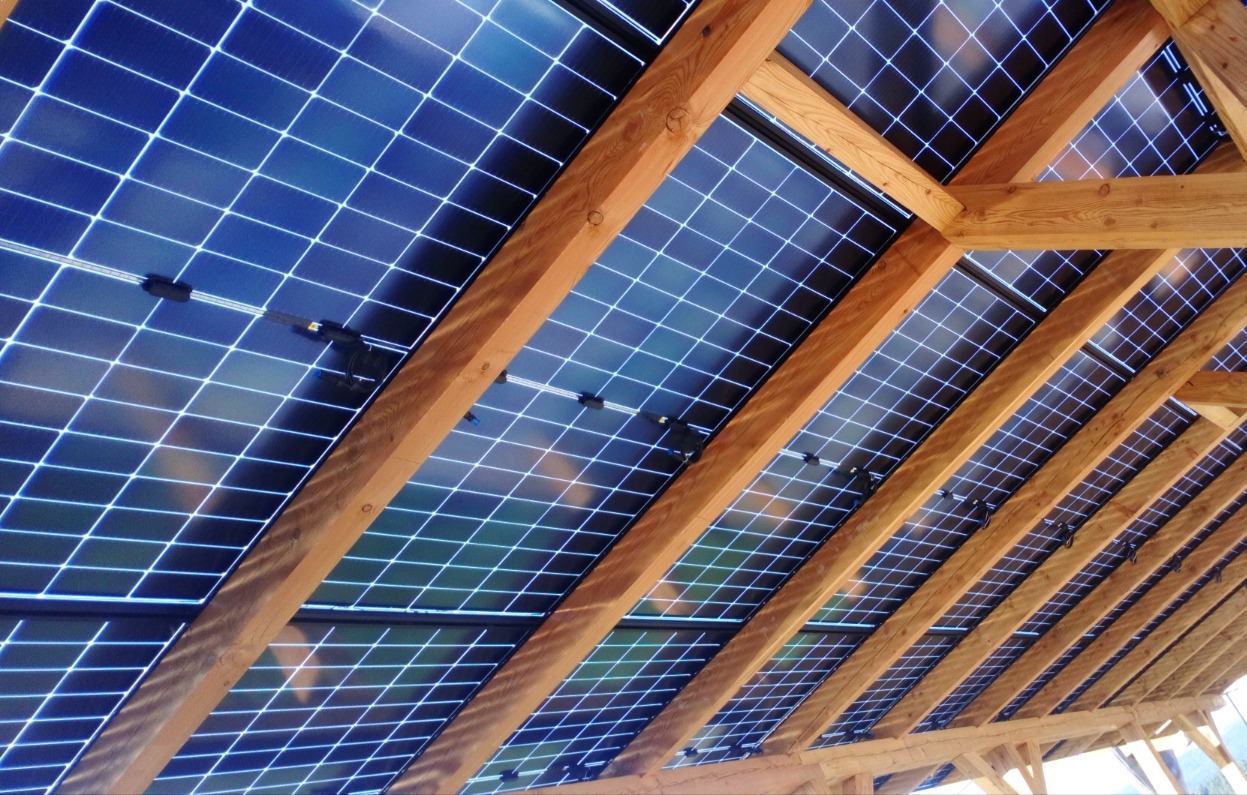 Widok od dołu na panele słoneczne, zainstalowane między drewnianymi belkami konstrukcji dachu, z widocznymi złączami kablowymi.