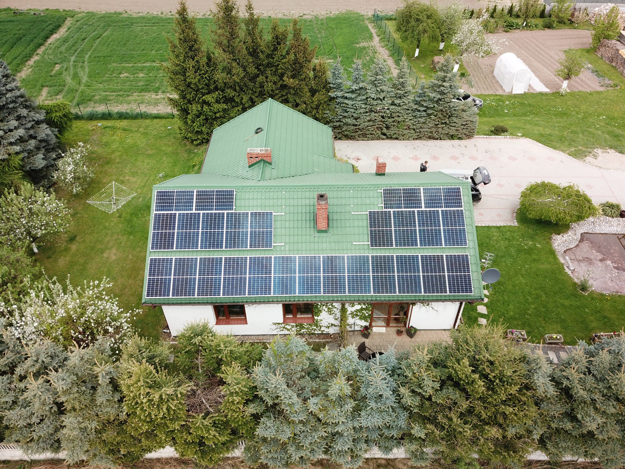 Widok z lotu ptaka na dom z zielonym dachem pokrytym panelami słonecznymi, otoczony zielenią i drzewami, z widocznym ogrodem i fragmentem drogi dojazdowej.
