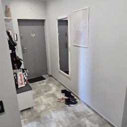 Wolski Wykończenia Wnętrz - Szare płytki podłogowe w przedpokoju z szarą ścianą, lustrem w białej ramie i grafiką. Widoczne buty i szafka na buty. Nowoczesne oświetlenie sufitowe.