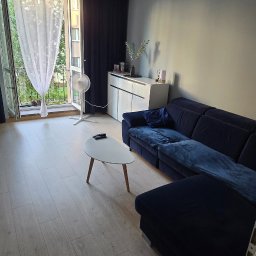 Wolski Wykończenia Wnętrz - Jasne panele podłogowe w salonie z granatową sofą narożną i białym stolikiem kawowym. Widok na balkon z firanką i zasłoną. Oświetlenie sufitowe punktowe.