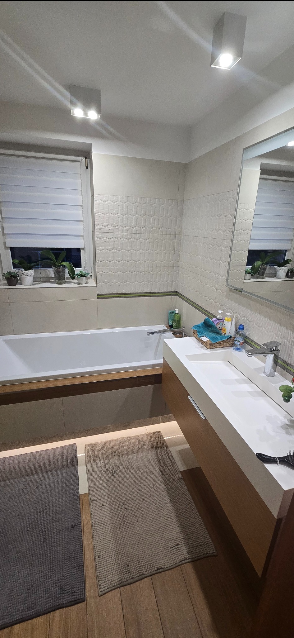 Jasna łazienka z wanną, umywalką z szafką i oknem z roletą. Na parapecie rośliny w doniczkach. Na podłodze dwa dywaniki. Oświetlenie sufitowe.