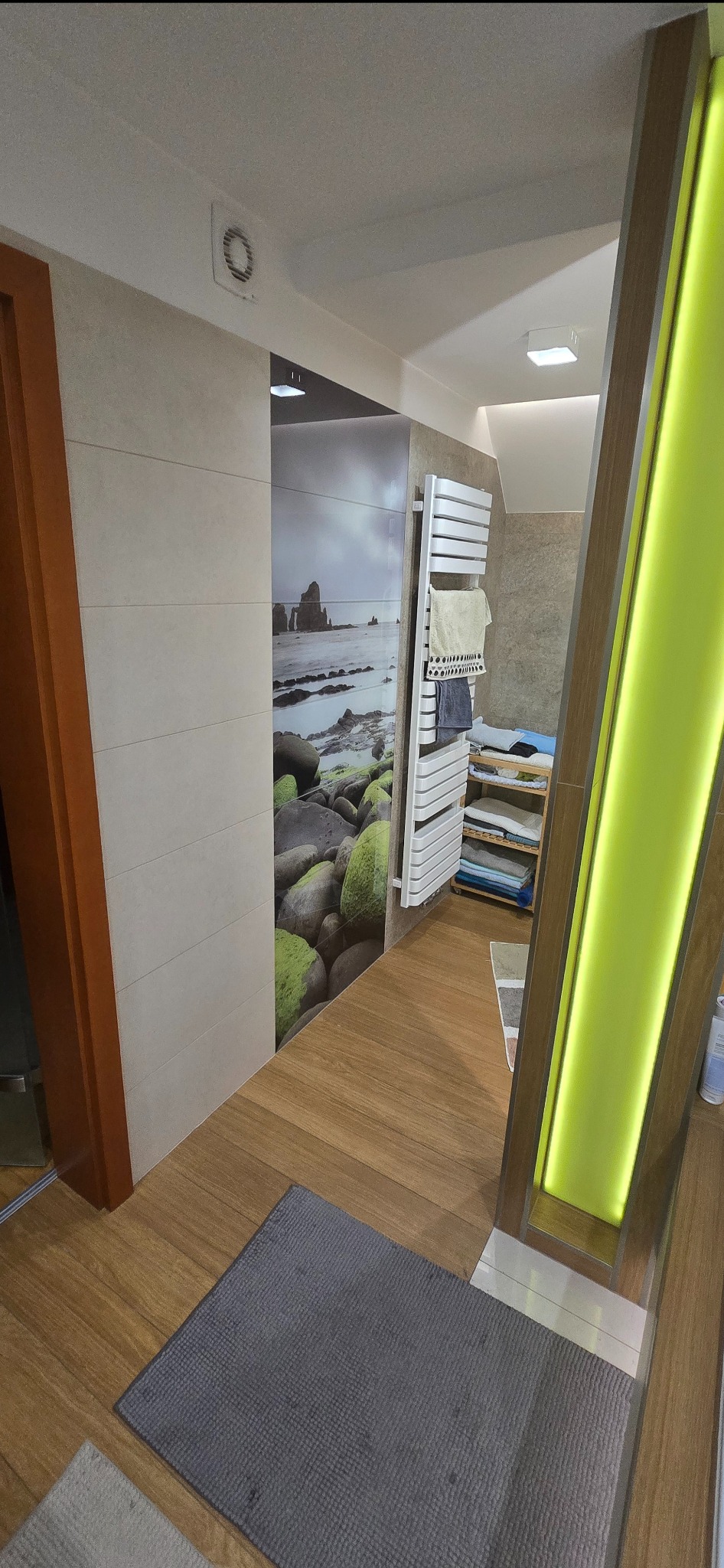 Wykończona łazienka z drewnianą podłogą, grzejnikiem drabinkowym i fototapetą z motywem morskim. Widoczny szary dywanik i oświetlenie LED.