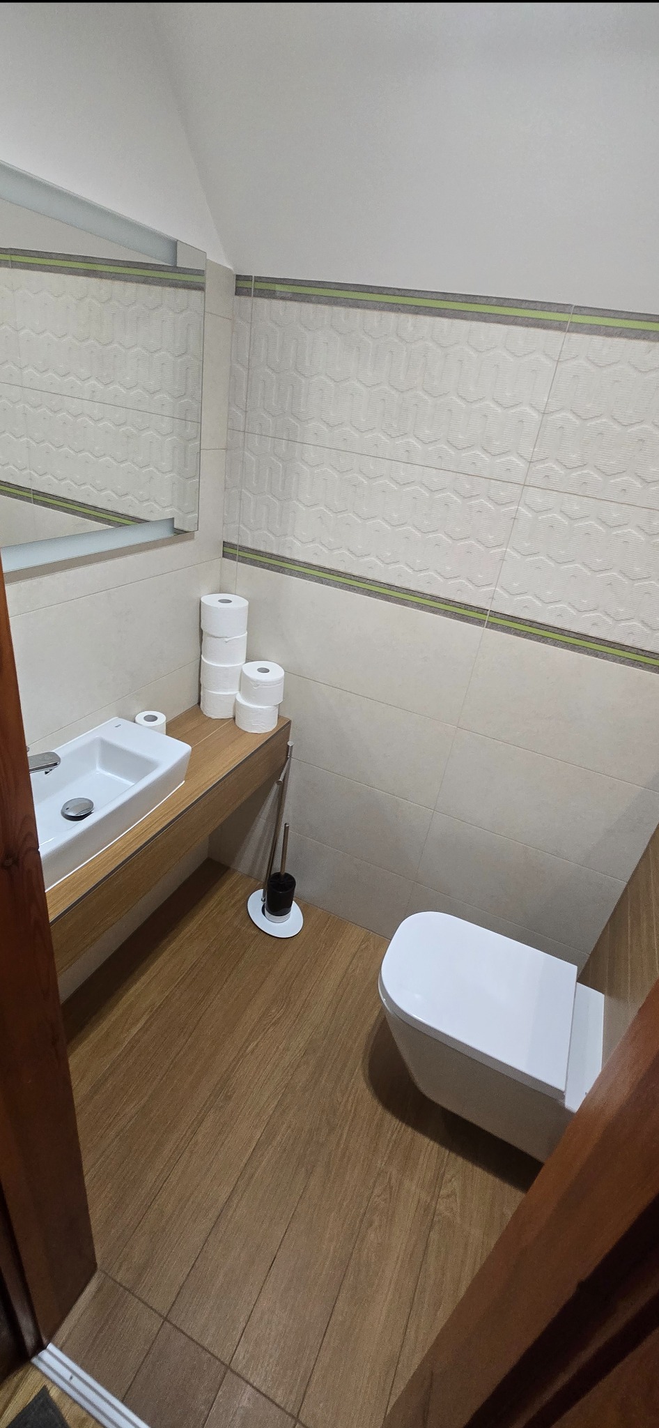 Jasna łazienka z drewnianą podłogą i blatem pod umywalką. Na blacie biała umywalka i stos rolek papieru toaletowego. Na ścianach białe płytki z delikatnym wzorem.