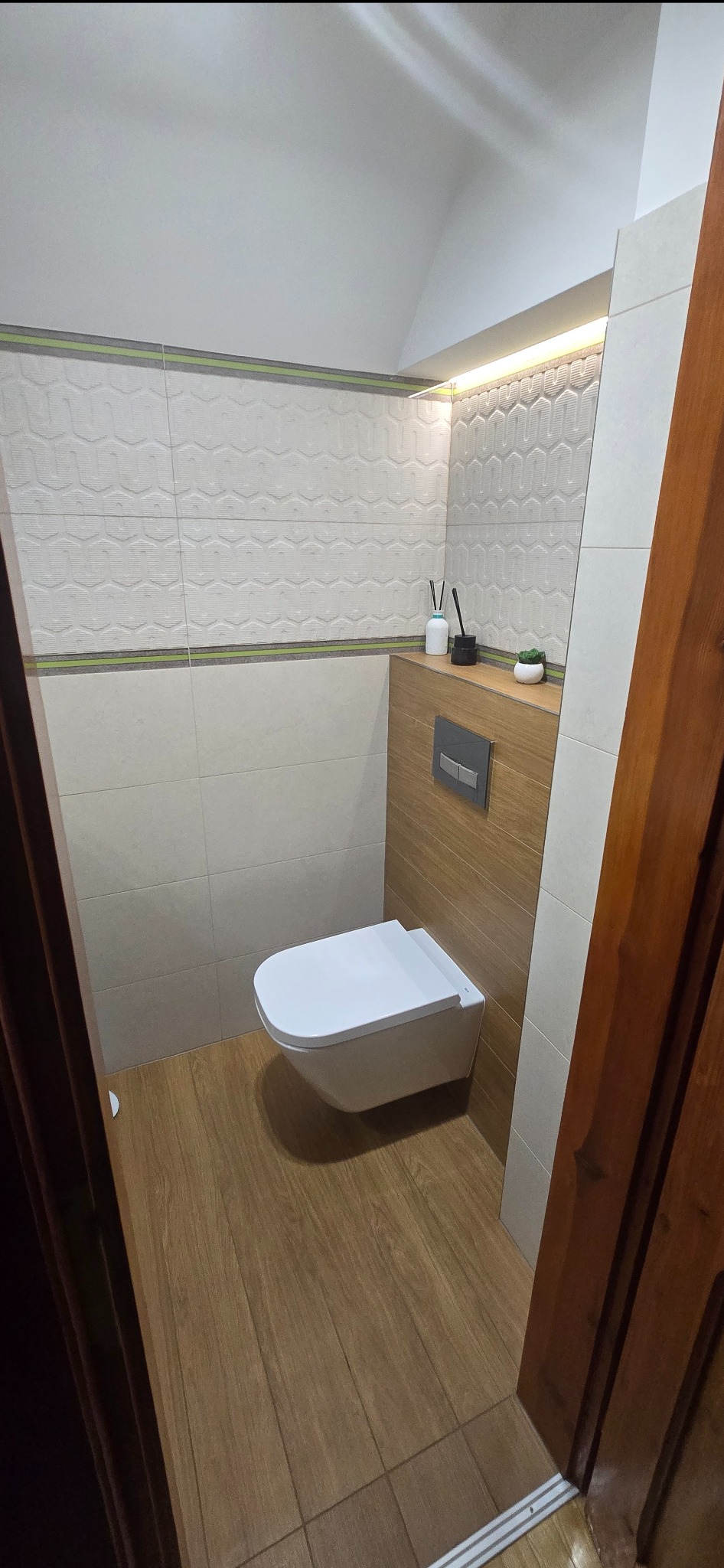 Nowoczesna toaleta podwieszana w wykończonej łazience. Drewnopodobne płytki na podłodze i ścianie, dekoracyjne płytki ścienne z poziomym paskiem. Oświetlenie LED nad zabudową.