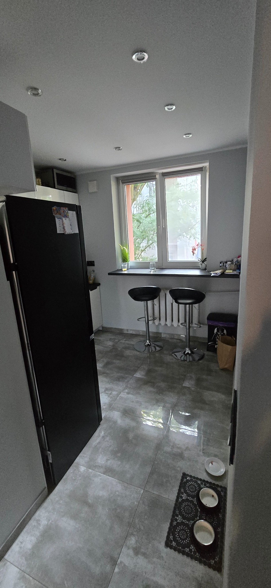 Jasna kuchnia z szarymi płytkami podłogowymi i czarną lodówką po lewej. Dwa hokery przy oknie. Miski dla zwierząt na podłodze. Minimalistyczny wystrój.