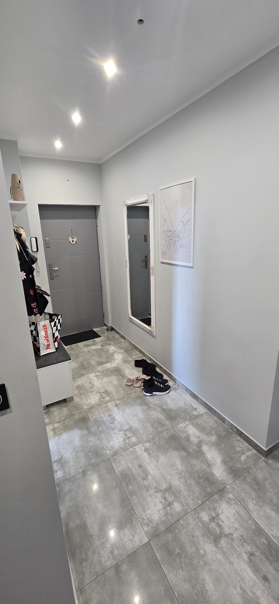 Szare płytki podłogowe w przedpokoju z szarą ścianą, lustrem w białej ramie i grafiką. Widoczne buty i szafka na buty. Nowoczesne oświetlenie sufitowe.