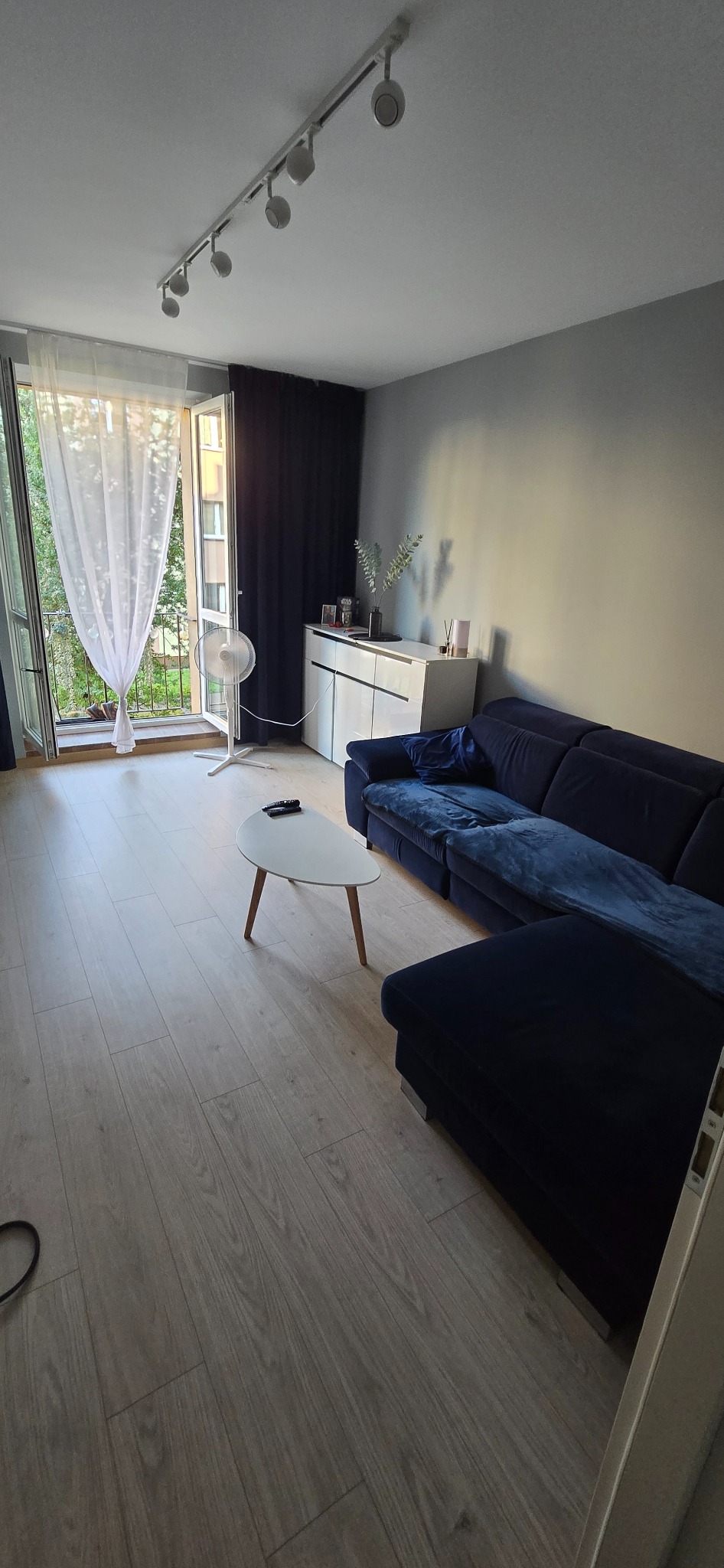 Jasne panele podłogowe w salonie z granatową sofą narożną i białym stolikiem kawowym. Widok na balkon z firanką i zasłoną. Oświetlenie sufitowe punktowe.