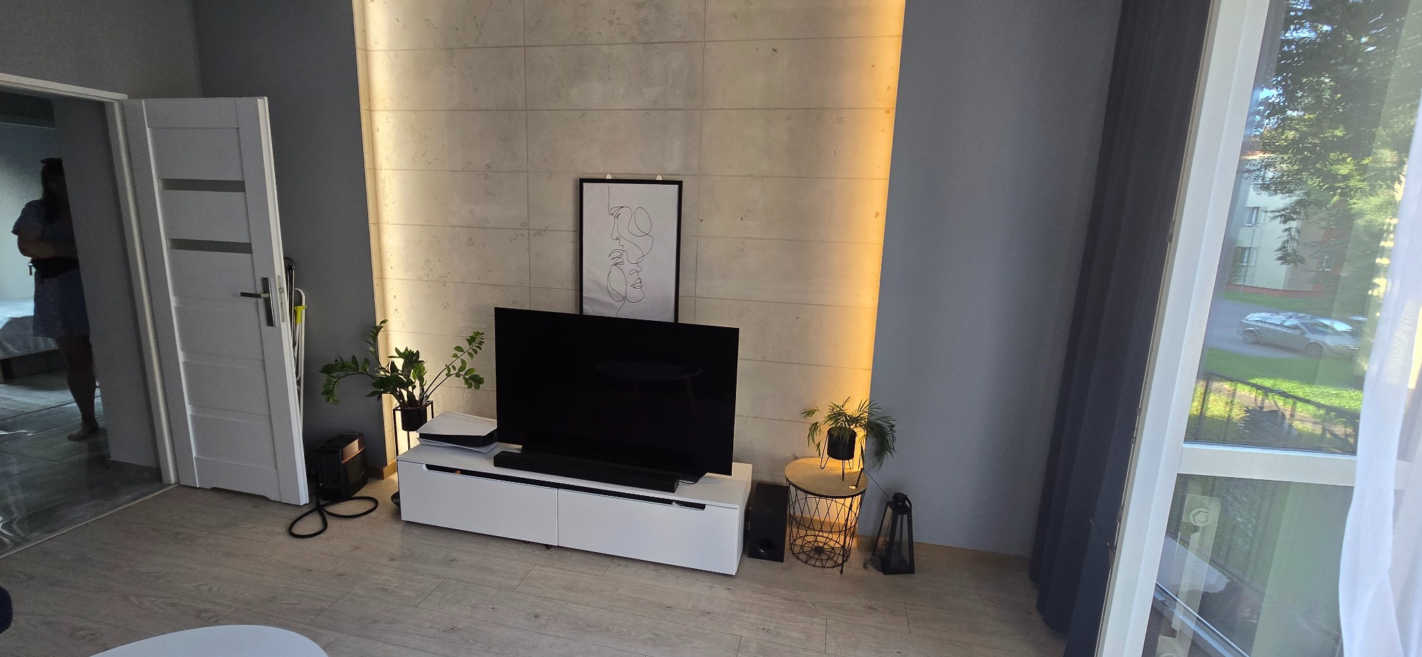 Nowoczesny salon z betonową ścianą podświetloną paskiem LED, białą szafką RTV i minimalistycznym plakatem. Widok na zieleń za oknem i fragment otwartych drzwi.