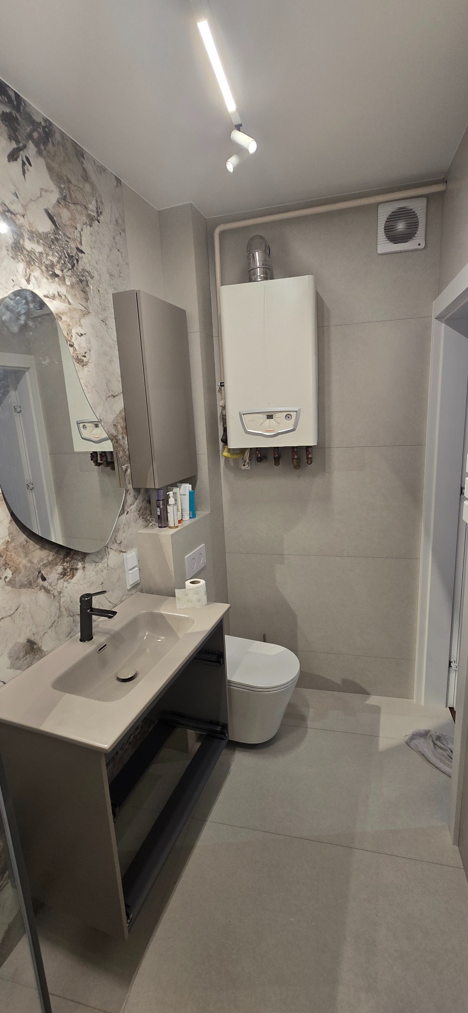 Nowoczesna łazienka po remoncie: szare płytki, umywalka z szafką, owalne lustro z podświetleniem, wisząca toaleta i piec gazowy. Minimalistyczny design.