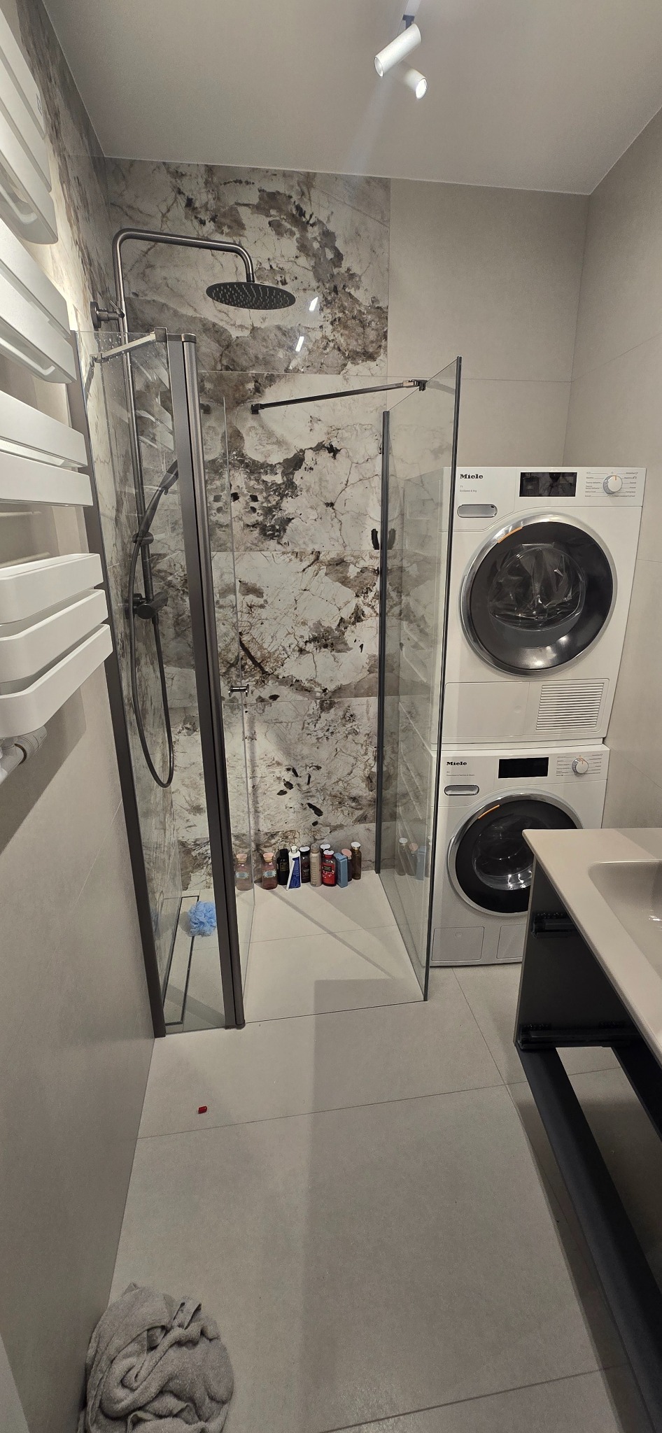 Wykończona łazienka z kabiną prysznicową typu walk-in, marmurowymi płytkami i pralką Miele. Nowoczesny design z szarymi płytkami podłogowymi i grzejnikiem drabinkowym.