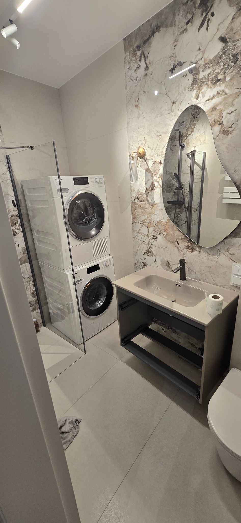 Wykończona łazienka z pralką i suszarką Miele w kolumnie, umywalką z szafką bez frontów i lustrem o nieregularnym kształcie na tle marmurowych płytek.