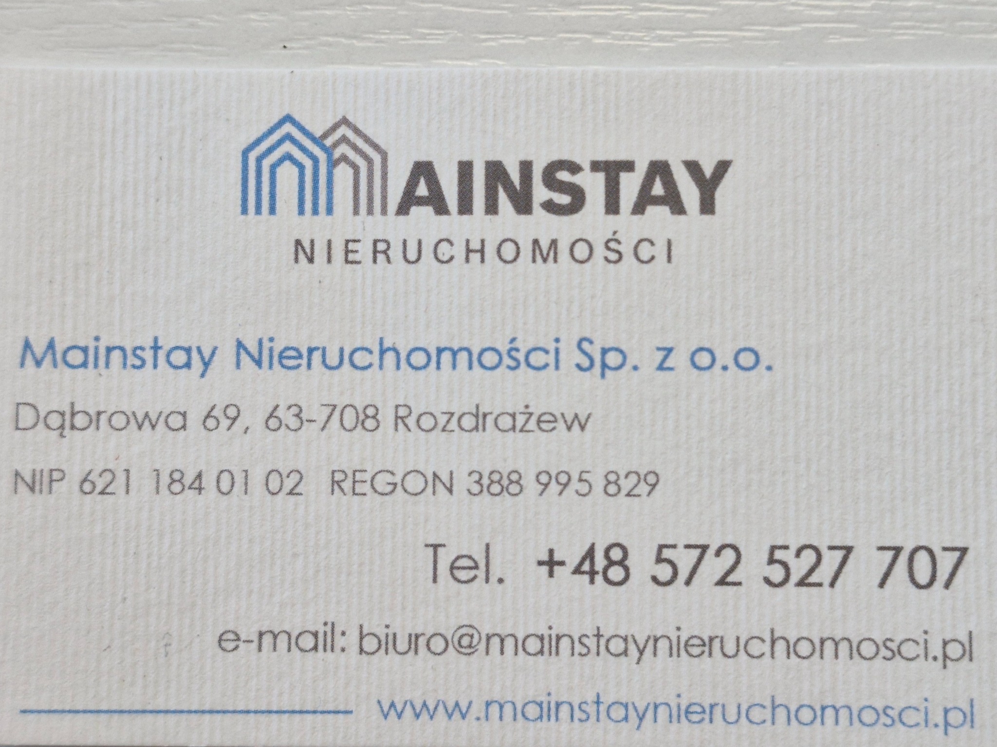 Wizytówka firmy Mainstay Nieruchomości z logo przedstawiającym schematyczny zarys dwóch połączonych domów w kolorze niebieskim i szarym, adres, NIP, REGON, numer telefonu i adres e-mail.