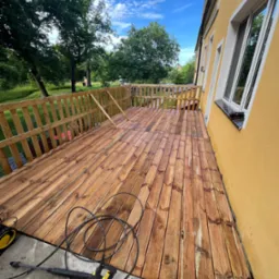 Świeżo wyczyszczony drewniany taras przydomowy z balustradą, z widoczną myjką ciśnieniową na betonowej posadzce, obok ż&oacute;łtej ściany budynku w słoneczny dzień.