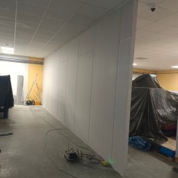 Damian - Montaż ścian działowych z płyt karton-gips w biurze. Widoczny laser krzyżowy, przewody i narzędzia. Część pomieszczenia zakryta folią malarską.