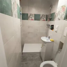 Wykończona łazienka w beżowej tonacji z akcentami kwiatowymi. Kabina prysznicowa z niskim brodzikiem, umywalka z szafką i toaleta. Na ścianach płytki imitujące kamień.