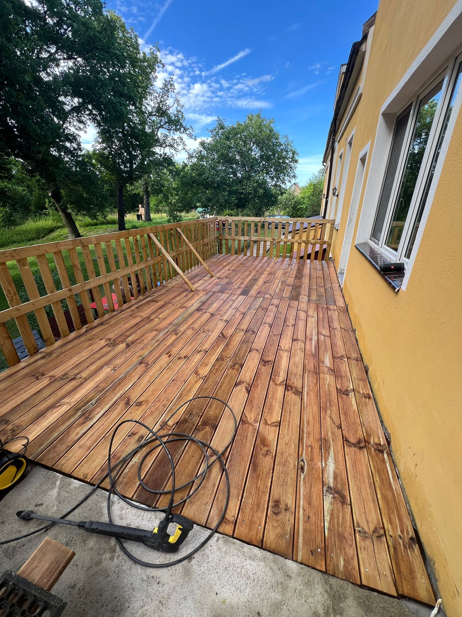 Świeżo wyczyszczony drewniany taras przydomowy z balustradą, z widoczną myjką ciśnieniową na betonowej posadzce, obok żółtej ściany budynku w słoneczny dzień.