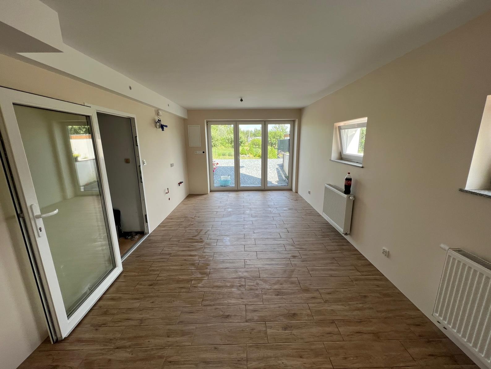 Salon po remoncie z drewnopodobną podłogą z płytek, białymi ścianami i grzejnikami. Duże okna balkonowe wpuszczają dużo światła. Widoczne przygotowania do wykończenia.